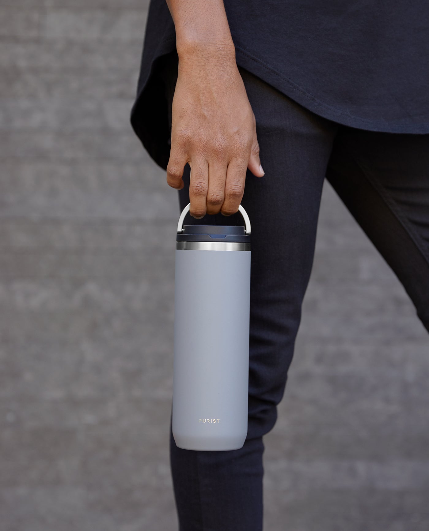 Shot of person walking and holding Purist Collective Mover bottle in ash with Element Lid by the handle.