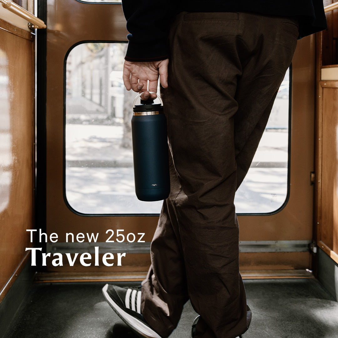 Person standing inside a train holding a sleek, dark blue 25oz Traveler bottle by the loop handle. Text reads 'The new 25oz Traveler.' The scene suggests a stylish, on-the-go lifestyle.