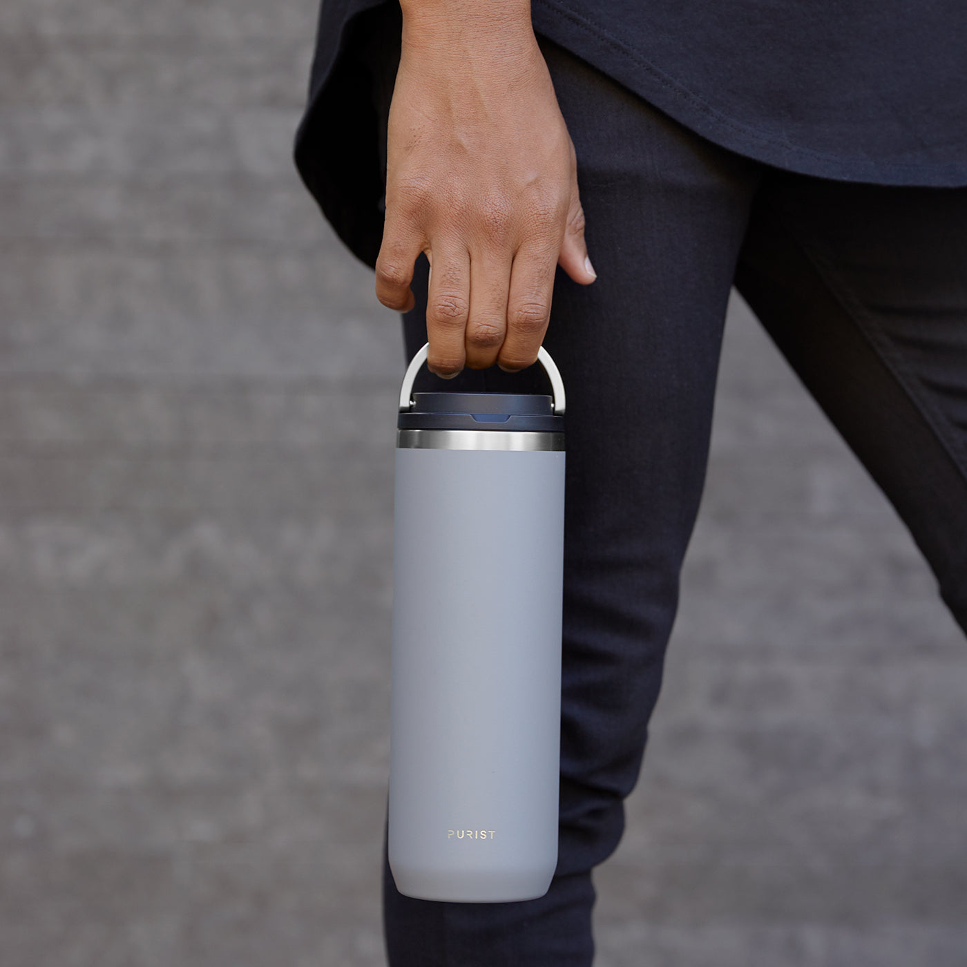Shot of person walking and holding Purist Collective Mover bottle in ash with Element Lid by the handle.